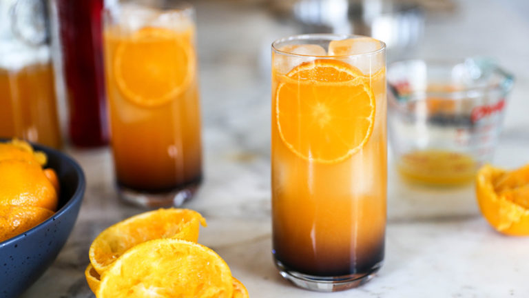 A photo of a mocktail recipe with a glass full of ice and orange liquid on a counter with several slices of oranges next to it