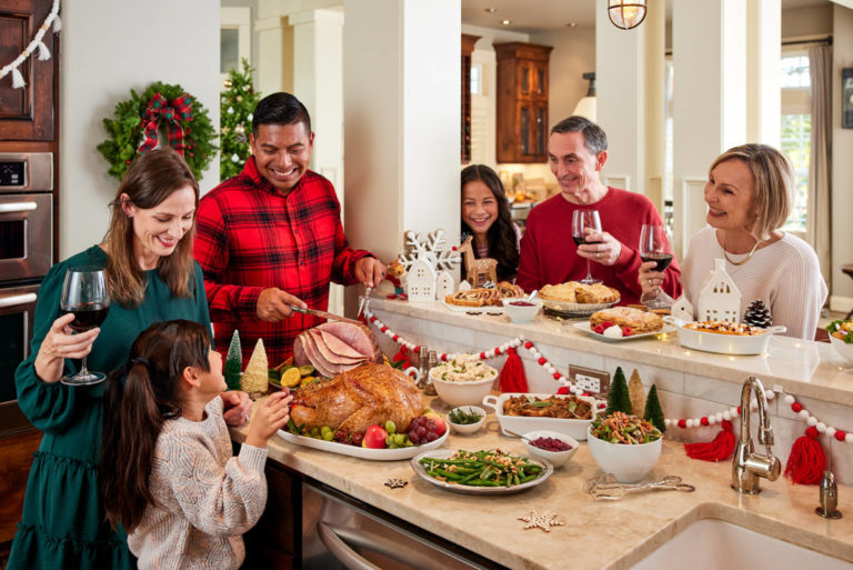 A photo of a holiday party with a family cutting roast turkey together in a holiday decorated kitchen
